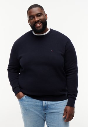 Uomo sorridente con barba che indossa un maglione blu navy e jeans azzurri chiari, in piedi con una mano in tasca su uno sfondo bianco.