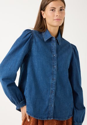 Frau trägt ein langärmeliges blaues Jeanshemd mit Knöpfen und gepufften Schultern, kombiniert mit braunen strukturierten Hosen.