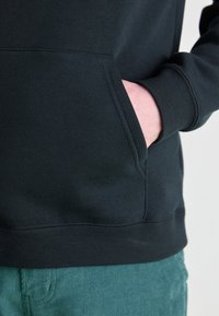 Sweatshirt noir avec une poche kangourou et des poignets côtelés, présenté sur un fond texturé clair, assorti à un pantalon en velours côtelé vert.