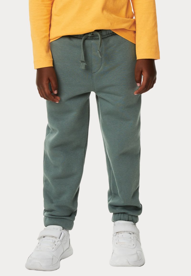 Grønne sweatpants med snoreliv, tapered cuffs og en blød tekstur, parret med hvide atletiske sko og en gul langærmet skjorte.