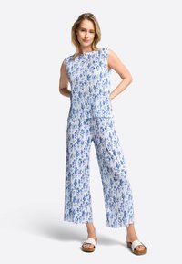 Blumenmuster-Jumpsuit in Blau und Weiß, leichtes Material, ärmelloser Oberteil, weit geschnittene Hosen, kombiniert mit weißen Sandalen.