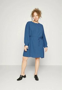 Vestido azul à altura do joelho com mangas longas e bufantes, decote redondo e um cinto amarrado na cintura. Usado com sapatos pretos e texturizados.