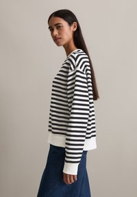 Frau mit langen dunklen Haaren, die ein schwarz-weiß gestreiftes Sweatshirt und einen dunkelblauen Jeansrock trägt, steht vor einem einfarbigen Hintergrund.