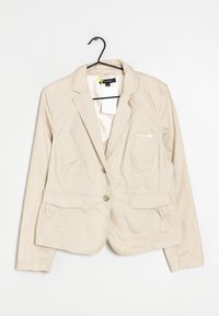 Blazer beige pour femme en tissu léger. Présente une fermeture à deux boutons, deux poches avant et une petite poche poitrine.