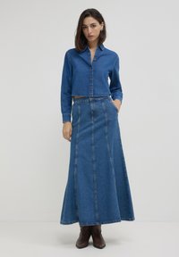 Femme portant une chemise en denim bleu à boutons et une longue jupe en denim assortie, avec des bottines en cuir brun, se tenant devant un fond blanc uni.