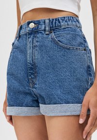 Jeansshorts med hög midja, mellanmörkblå tvätt, uppvikta fållar, synliga sömmar och knappstängning framtill. Har sidfickor.