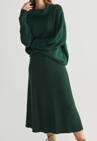 Kobieta ubrana w ciemnozieloną spódnicę użytą z prążkowanej dzianiny oraz dopasowany luźny sweter z dekoltem w wodę, w połączeniu z beżowymi butami na sznurówki.