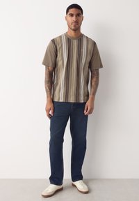 Földszínű rövid ujjú póló grafikai mintával, változó szélességű függőleges csíkokkal, sötétkék farmerrel és fehér tornacipővel párosítva.