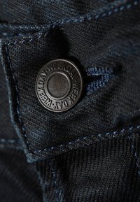 Gros plan sur un tissu en denim foncé avec des coutures bleues et un bouton en métal noir gravé du mot « WEEKDAY » répété tout autour du bord.