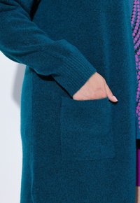 Teal stickad kappa har ribbade ärmslut, sidoficka och textiler, vilket ger ett mjukt och varmt utseende.