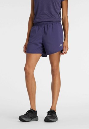 Person iført mørkeblå New Balance atletikshorts, mørkeblå t-shirt og sorte løbesko, stående mod en ensfarvet baggrund.