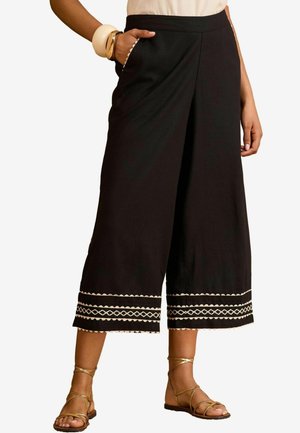 Culottes negre dintr-un material moale, cu broderie decorativă de culoare crem la tiv și buzunare laterale. Acestea sunt acompaniate de sandale cu bretele.