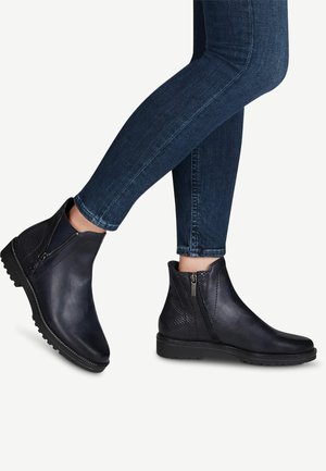 Personne portant des bottines en cuir bleu foncé avec fermetures éclair latérales et panneaux texturés à l'arrière, associées à un jean bleu foncé ajusté.