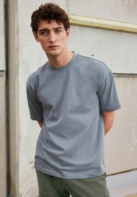 Tricou gri din bumbac, cu mâneci scurte și guler rotund, purtat cu pantaloni verzi, pe un fundal de zid de beton.