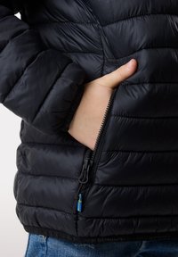 Giubbotto imbottito nero con un design trapuntato, mano in una tasca laterale, presenta una chiusura con zip e un accento di tessuto blu e giallo sulla zip.