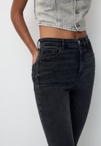 Femme portant un jean noir taille haute et un gilet en denim gris clair cropped avec des boutons, la main dans la poche, sur un fond uni.