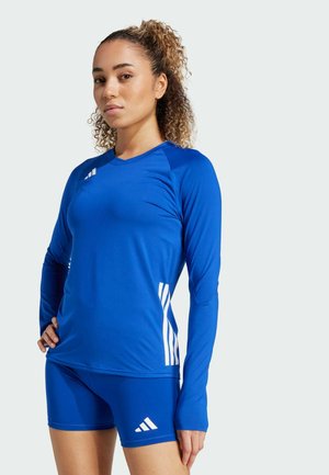 Młoda kobieta o kręconych włosach ubrana w niebieską sportową bluzkę z długim rękawem oraz dopasowane spodenki z białymi logo Adidas.
