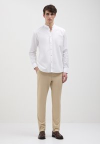 Camicia bianca a maniche lunghe con colletto alla cinese, abbottonatura frontale, e pantaloni beige, indossata con scarpe marroni su uno sfondo neutro.