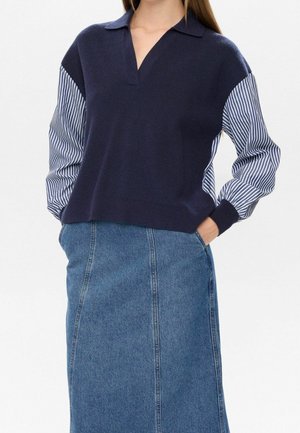 Femme portant un pull bleu marine à col avec des manches rayées bleu et blanc et une jupe en jean bleue, les mains dans les poches de la jupe.