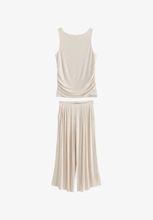 Vrouw met krullend haar en een beige mouwloze top en wijd uitlopende broek die op één voet staat naast gestapelde gestreepte kussens.