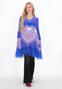 Poncho albastru, cu model, având un tiv cu franje, cu desene geometrice în roz și alb. Textură moale și pufoasă. Asortat cu pantaloni negri.