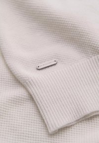 Bílý texturovaný svetr s hladkým ribovým lemem a malou stříbrnou visačkou připevněnou na látce.