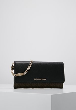 Zwarte en bruine Michael Kors portemonnee clutch met gouden kettingriem en logo op een wit piedestal tegen een grijze achtergrond.