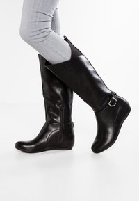 Bottes noires jusqu'au genou en cuir synthétique lisse, avec une fermeture éclair sur le côté, un accent de sangle et un bout rond. Semelle en caoutchouc avec un talon bas.