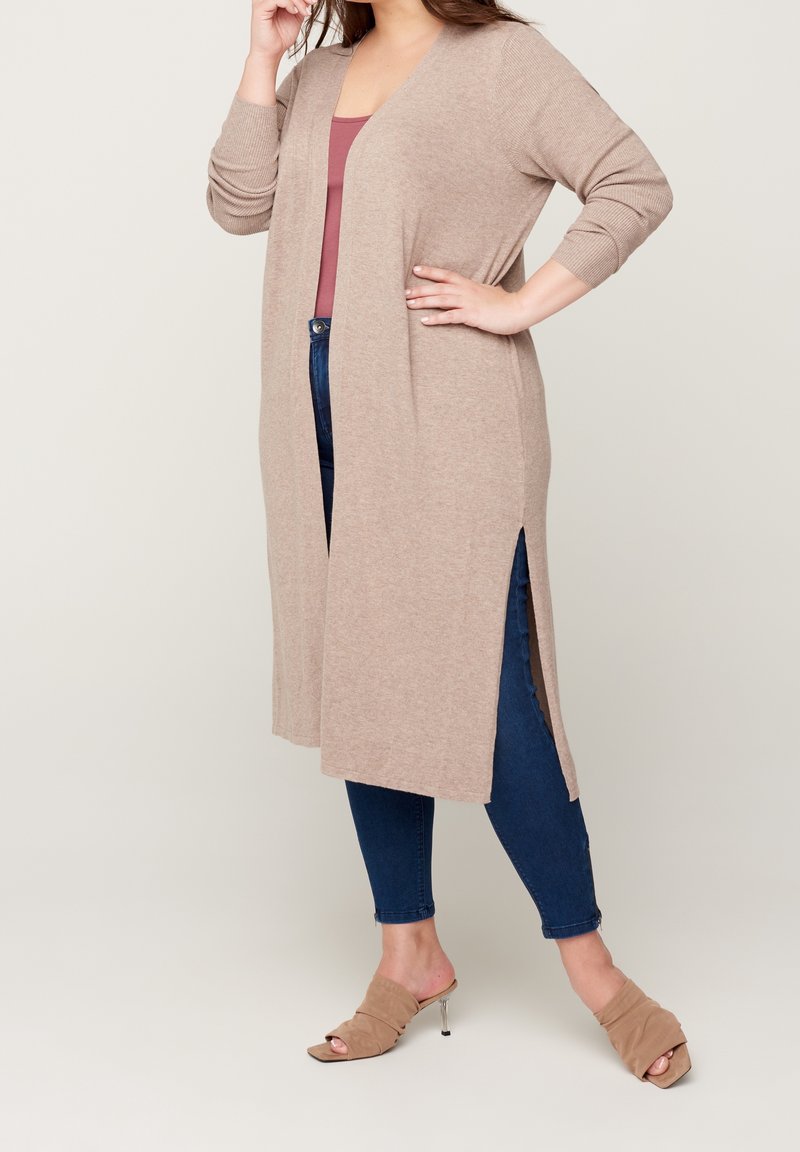 Femme portant un long cardigan beige avec des fentes latérales, un haut mauve, un jean skinny bleu, et des mules à talons ouvertes beiges, debout avec une main sur la hanche.