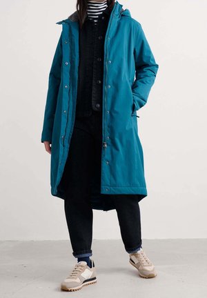 Lang teal hættetrøje jakke båret over sorte bukser, stribet højhalset trøje og sneakers, stående med den ene hånd i lommen på en lys gulv.