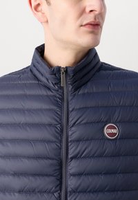 Jachetă izolată de culoare albastru închis, cu design matlasat pe orizontală, guler înalt și fermoar frontal. Prezintă un patch cu logo rotund pe pieptul stâng.