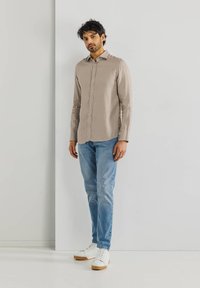 Hellbraunes Langarmhemd mit Knopfleiste, kombiniert mit blauen Jeans und weißen Sneakers mit Gummisohle.