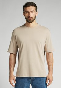Beige tričko s krátkým rukávem vyrobené z měkké, lehké tkaniny. Má kulatý výstřih a volný střih, v kombinaci s modrými džíny.