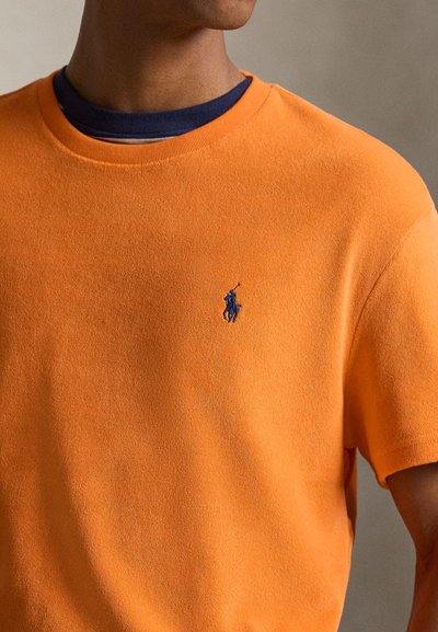 Άνδρας που φορά φωτεινό πορτοκαλί κοντομάνικο πουκάμισο με μπλε κεντημένο λογότυπο ποδηλάτη πόλο και σκούρα μπλε μπλουζάκι με στρογγυλή λαιμόκοψη από μέσα.