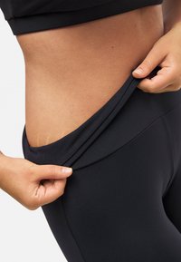 Leggings atléticos negros con una tela suave y elástica. Una mano está ajustando la cinturilla, revelando una textura blanda y elástica.