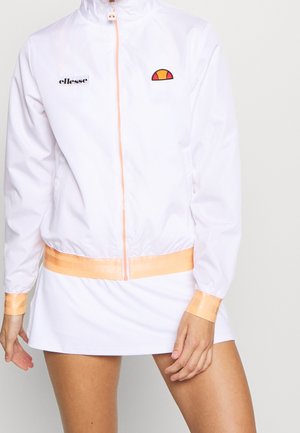 Witte jas met rits, oranje bies en logo's, gecombineerd met een witte rok, gedragen door een persoon met één hand ontspannen langs het lichaam.