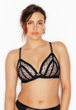 Femme portant un soutien-gorge en maille noire transparente avec des motifs géométriques, des boucles d'oreilles en or en forme d'anneau, et les cheveux lâches et ondulés sur un fond blanc.