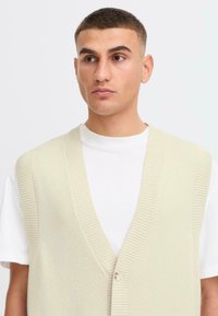 Un giovane con i capelli corti indossa un gilet beige chiaro su una camicia bianca, guardando leggermente di lato su uno sfondo semplice.