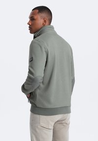 Fleece bunda v tlumené zelené barvě s vysokým límcem, strukturovanou tkaninou, šedými loketními nášivkami a zipovou kapsou na boku.