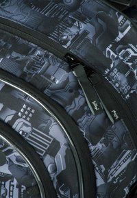 Zaino nero con un motivo geometrico in grigio e nero, dotato di doppie cerniere, tessuto testurizzato e cuciture rinforzate.