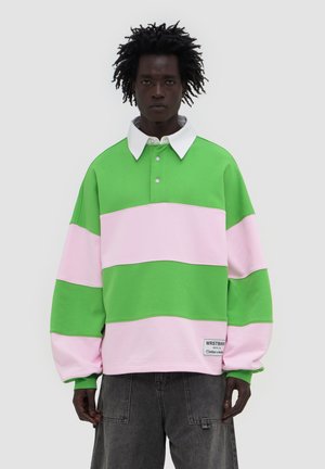 Homme portant un maillot de rugby vert et rose à rayures oversize avec un col blanc et un pantalon gris foncé, debout devant un fond uni.