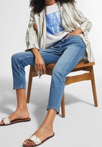 Blazer a rayas, camiseta gráfica blanca, jeans de mezclilla azul claro y sandalias blancas tipo zueco con un acento circular, sentado en una silla de madera.