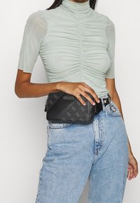 Schwarze Schultertasche mit Struktur und Logo-Muster, kombiniert mit einem hellgrünen enganliegenden Top und hellblauen, hochtaillierten Jeans.