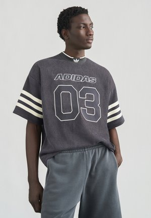 Nadcenené tričko Adidas v tmavosivej farbe s bielymi pruhmi, s rebrovanou textúrou, výraznou potlačou "03" a logom na golieri. Kombinované so sivými šortkami.