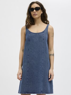 Femme portant une robe en jean bleu sans manches et des lunettes de soleil noires, debout devant un fond clair uni.