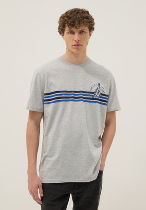 Junger Mann trägt ein graues T-Shirt mit blauen horizontalen Streifen und einem "Bella Vita"-Aufdruck auf der Brust, kombiniert mit schwarzen Hosen.