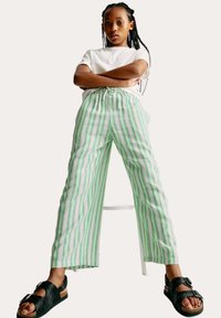 Gestreepte groene en witte lichte broek met een trekkoord in de taille, gecombineerd met een effen wit T-shirt met korte mouwen en zwarte sandalen.