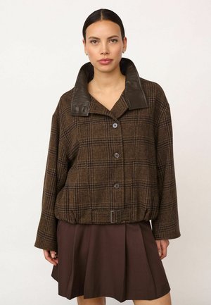 Femme portant une veste à carreaux marron boutonnée avec un col en cuir et une jupe plissée marron foncé, debout devant un fond uni.
