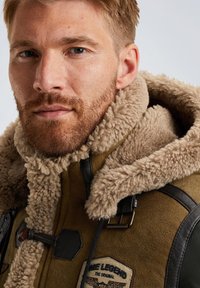 Bruine shearling jas met een fluffy kraag, leren accenten, een patch op de borst en een glad oppervlak. Gelaagd en warme uitstraling.