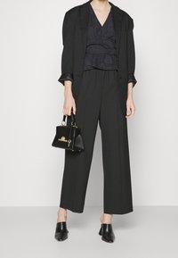 Mustassa räätälöidyssä bleiserissä mustan teksturoidun paidan päällä, leveälahkeiset housut ja teräväkärkiset mustat korkokengät. Pieni musta käsilaukku tuo yksityiskohtia.